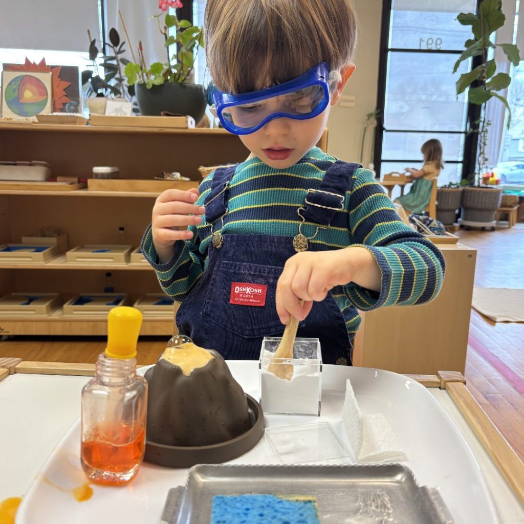 A Children's House student wearing safety goggles pours liquid into a model volcano