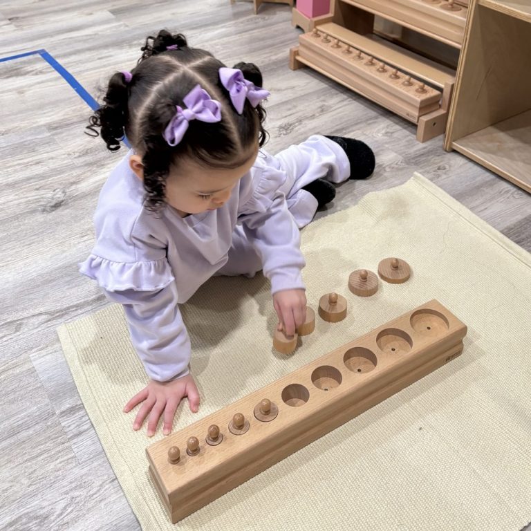 A toddler places knobbed cylinders into a wooden block on a work rug