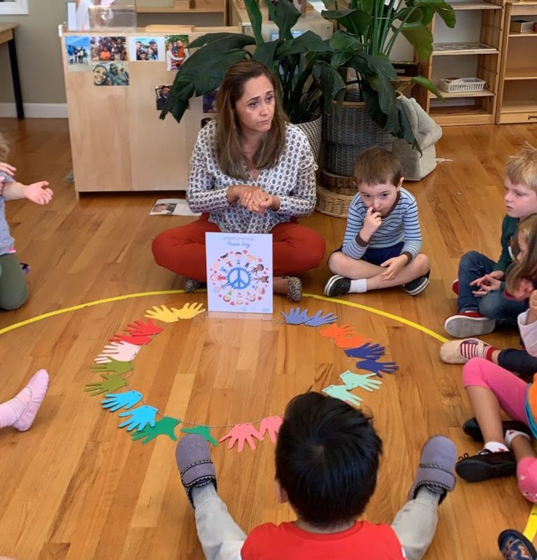 A guide sits with children in a circle around a ring of colorful paper hands during morning circle