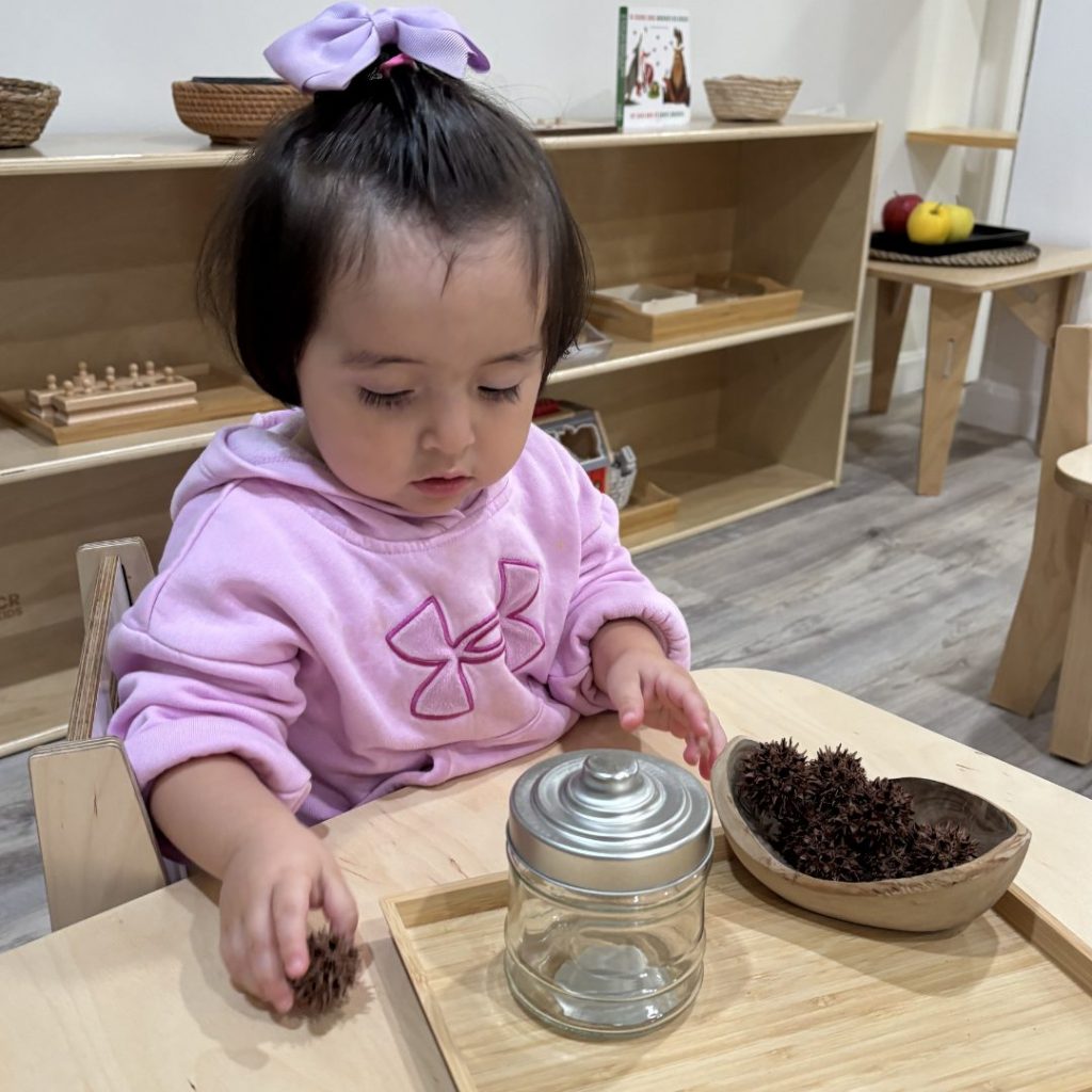 A toddler in the Nido community explores a sensory tray with a wooden scoop and glass jar