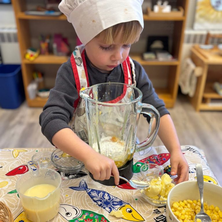 A child in a chef's hat and apron prepares food at a classroom table