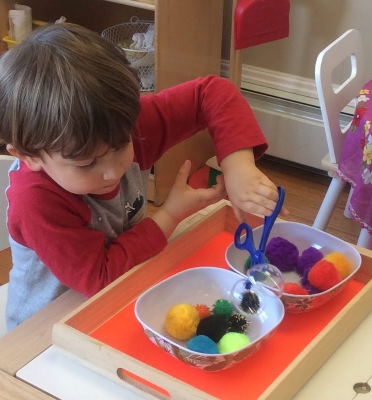 A child uses tongs to move colorful pom-poms between bowls on a tray